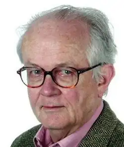 Colour three-quarter profile publicity shot, of clean-shaven, bespectacled half-smiling man, with receding short white hair, brushed back