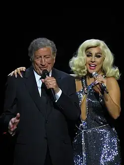 Bennett in suit on the left and Gaga in a sparkling dress and blonde hair singing onstage
