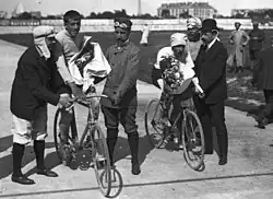 Two men with flowers in their hands, sitting on bicycles being held by other men, with the Eiffel tower seen in the background.