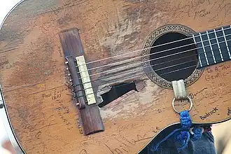 A classical guitar. There are several damages in the soundboard, near the sound hole there is a big hole and the wood is worn out in the surrounding areas of it. The guitar has several signatures on it. there is a blue and white strap in the soundhole.