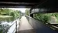 Looking westwards from the canal towpath at the "Three Bridges", showing the Brentford Branch Line in a cutting, and the underside of the road bridge above. Picture taken May 2025.