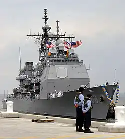 Color photo of a Navy cruiser docked alongside a concrete pier