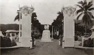 Two white pillars are topped with sculptures of bull's heads. A statue of a man holding a domestic animal can be seen beyond the pillars.