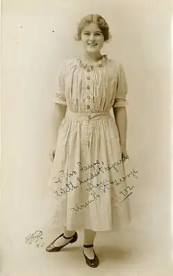 A young smiling white woman, standing, wearing a light-colored dress with buttons down the front of the bodice and a ruffled neckline.