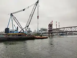 A large crane on a barge, about to lift the triangular metal frame supporting 3 turbines