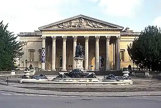 A Palladian style nineteenth century stone building with a large colonnaded porch. In front a large metal statue on a pedestal and fountains with decorations.