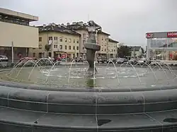 A fountain in a square in Gospić, Croatia. The centrepiece is a stone statue with a bust of a nude woman, only one arm intact, atop a jagged pedestal. In the pool around it is a circle of jets which spray water inwards toward the statue.