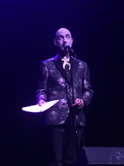 Bald white man wearing a skull and rose patterned suit, standing in front of a microphone holding a script, in a theatre with purple lighting.