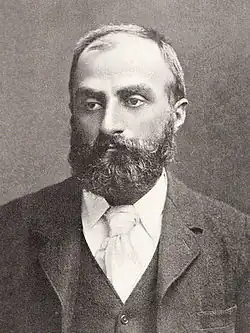 Heavily bearded man with dark receding hair, wearing a dark coloured jacket, white-collar and pale tie. He is looking slightly to the left, with a solemn expression