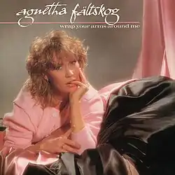 A young woman poses in front of a background full of pink and black satin fabrics. She is wearing a pink buttoned-up sweater, resting her chin on her hand, with her left hand pinky finger in her mouth. To the top left, the text is written in elegant style, and it reads "Agnetha Fältskog", with a pink underline. The bottom right of the underline shows a text all in lower-case reading "Wrap Your Arms Around Me".