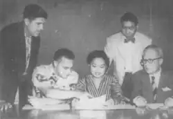 Photograph of a young woman looking at a paper, surrounded by four men.