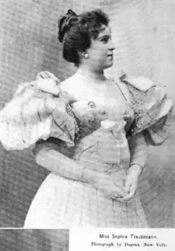 A young white woman with dark hair in a bouffant updo; she is standing, hands together in front of her midsection; she is wearing a light-colored gown with very puffy round sleeves and a low square neckline